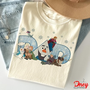 May include: Cream-colored t-shirt with the word "DAD" in blue, featuring characters from the movie Frozen. The design includes Olaf, Elsa, Anna, Kristoff, Sven, and snowflakes. The shirt is folded on a pair of blue jeans.