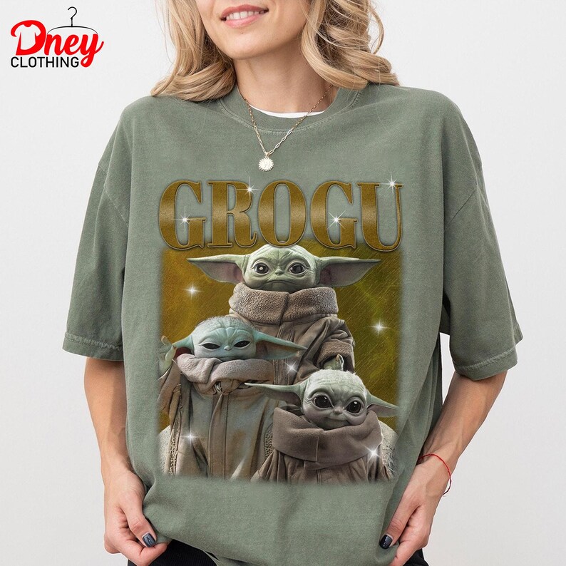 K&ouml;nnte beinhalten: Olivgr&uuml;nes T-Shirt mit einem Aufdruck von Grogu aus The Mandalorian. Das Wort "GROGU" ist in goldenen Buchstaben &uuml;ber dem Bild von Grogu aufgedruckt. Das Bild zeigt drei Grogu-Charaktere in braunen Roben.