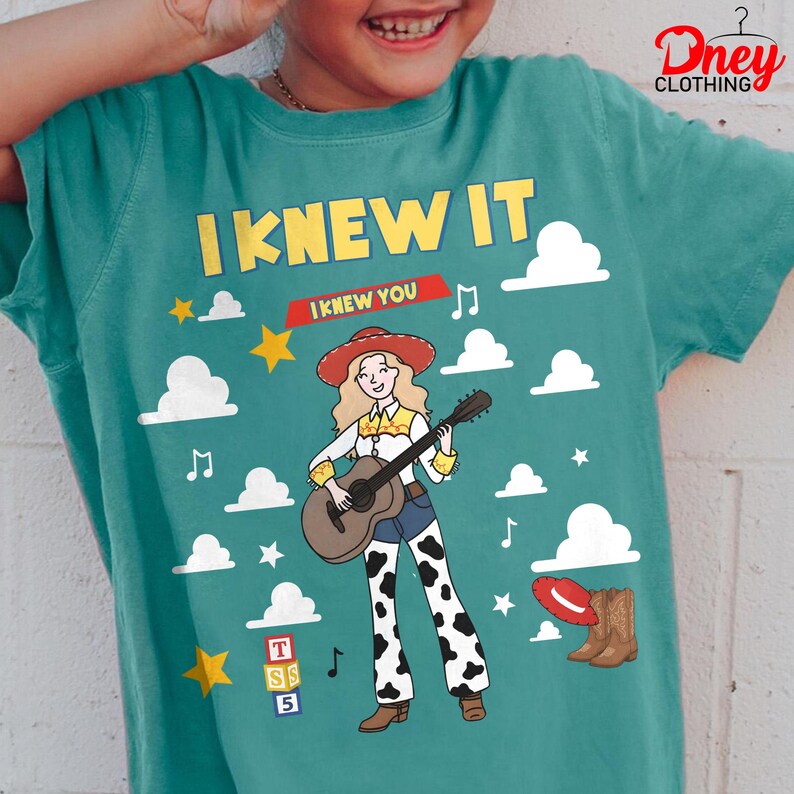 May include: A teal t-shirt featuring a graphic of a cowgirl with blonde hair playing an acoustic guitar. The design includes the text I KNEW IT and I KNEW YOU, white clouds, yellow stars, musical notes, a red hat, brown boots, and alphabet blocks.