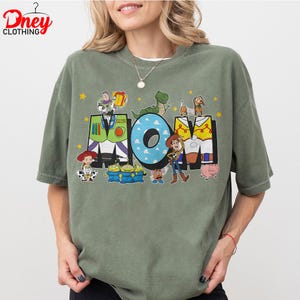May include: Sage green t-shirt with a graphic design featuring the word "MOM" in a playful font, decorated with characters from the Toy Story film. The shirt has short sleeves and a crew neck.