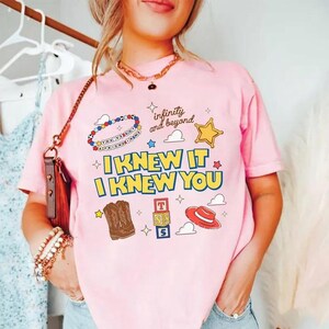 Puede incluir: Una camiseta de manga corta de color rosa claro con un estampado gr&aacute;fico que incluye el texto "I KNEW IT I KNEW YOU", "infinity and beyond" y "YOU'VE GOT A FRIEND IN ME". El dise&ntilde;o incluye ilustraciones de una bota vaquera, un sombrero rojo y bloques.