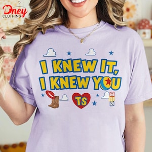 Puede incluir: Una camiseta de color morado claro con el texto en amarillo y azul I KNEW IT, I KNEW YOU. El gr&aacute;fico incluye un sombrero de vaquero rojo, botas marrones, un coraz&oacute;n rojo con las letras TS, una pelota azul y amarilla y bloques de juguete.
