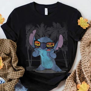Op de afbeelding: Een zwart t-shirt met korte mouwen en een afbeelding van een blauw alien-personage met een zonnebril waarin een zonsondergang en palmbomen weerspiegelen. Het personage heeft grote oren, steekt de tong uit en staat voor palmbomen.