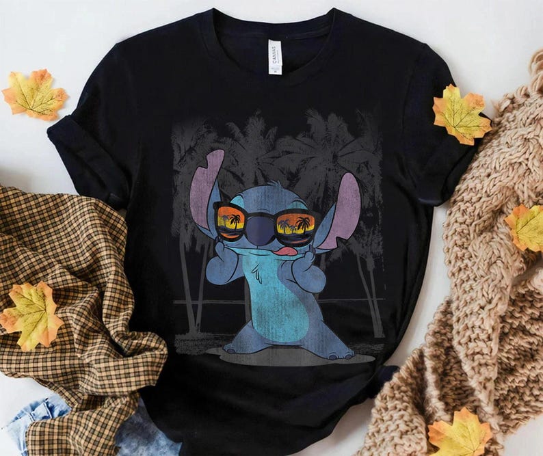 Op de afbeelding: Een zwart t-shirt met korte mouwen en een afbeelding van een blauw alien-personage met een zonnebril waarin een zonsondergang en palmbomen weerspiegelen. Het personage heeft grote oren, steekt de tong uit en staat voor palmbomen.