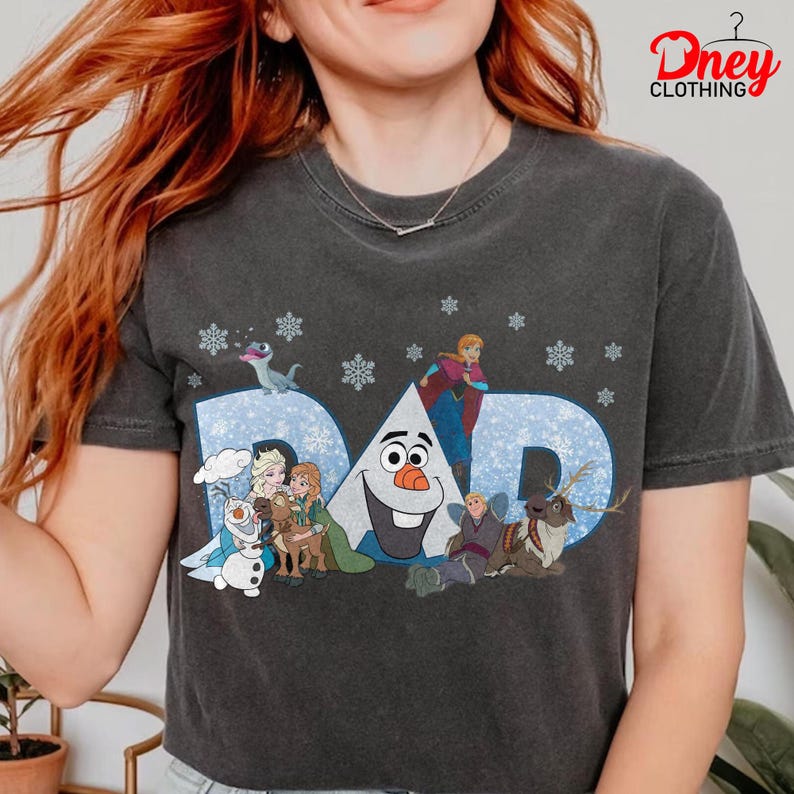 May include: Dark gray t-shirt featuring the word "DAD" in light blue, with snowflakes and characters from a popular animated movie. The shirt has a crew neck and short sleeves.
