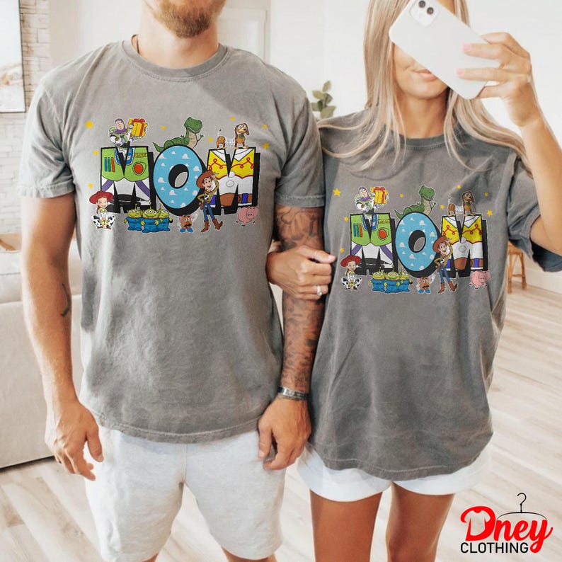 May include: Two grey t-shirts featuring the word "MOM" in large, colourful letters. The letters are decorated with cartoon characters. The shirts are short-sleeved and have a vintage wash appearance.