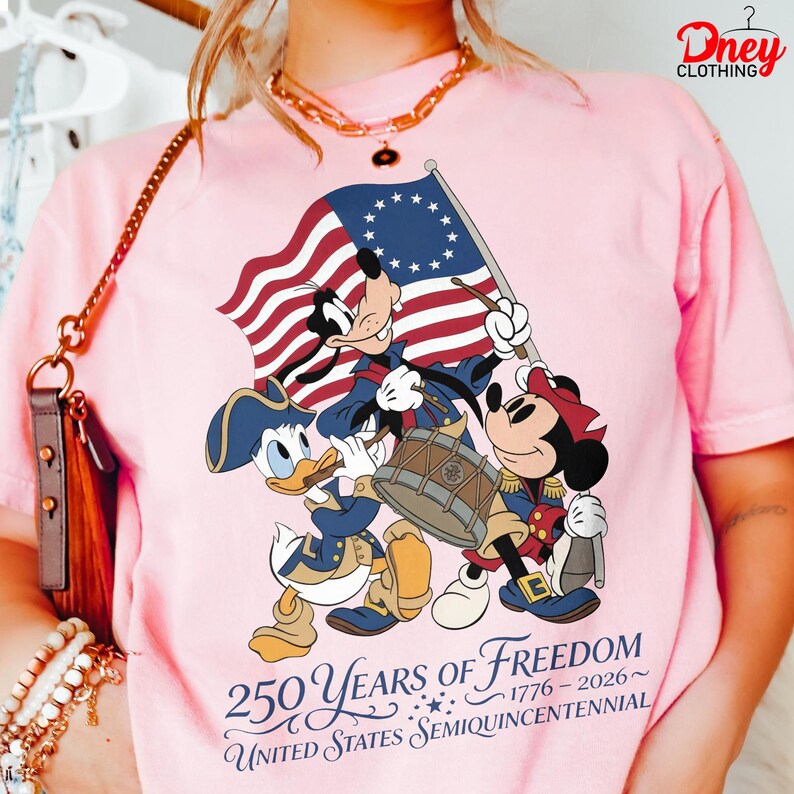 May include: Pale pink t-shirt with a patriotic design featuring Disney characters Goofy, Donald Duck, and Mickey Mouse in colonial outfits. The design includes a flag and the text "250 Years of Freedom 1776-2026 United States Semiquincentennial."