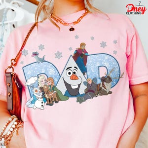 May include: Pink t-shirt featuring the word "DAD" in blue with a snowflake pattern. The shirt has cartoon characters from the movie Frozen, including Olaf, Anna, Elsa, Kristoff, Sven, and Bruni. The shirt has the "Dney Clothing" logo.