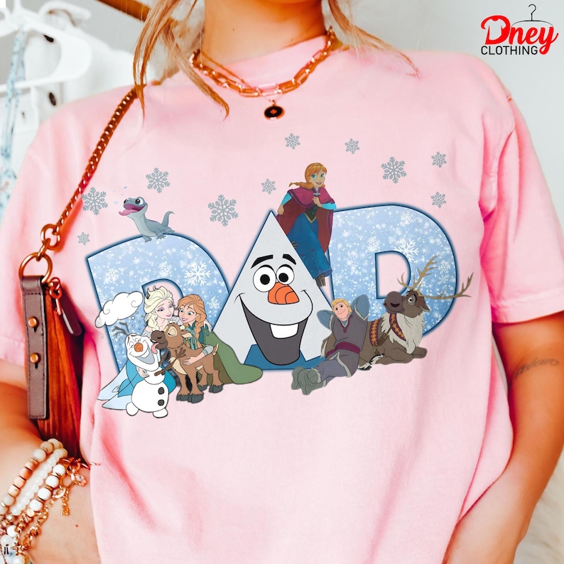 May include: Pink t-shirt featuring the word "DAD" in blue with a snowflake pattern. The shirt has cartoon characters from the movie Frozen, including Olaf, Anna, Elsa, Kristoff, Sven, and Bruni. The shirt has the "Dney Clothing" logo.