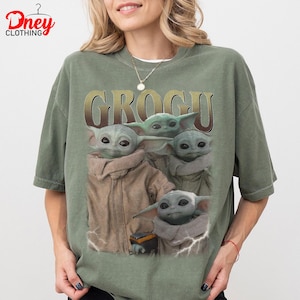 K&ouml;nnte beinhalten: Ein olivgr&uuml;nes T-Shirt mit dem Wort "GROGU" in goldfarbenen Buchstaben. Das Shirt zeigt eine Grafik von Grogu, auch bekannt als Baby Yoda, aus der Star Wars-Serie. Die Grafik zeigt Grogu in verschiedenen Posen.