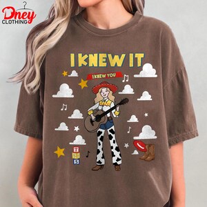 May include: A brown short-sleeved t-shirt featuring a graphic of a cowgirl playing a guitar. The text reads I KNEW IT and I KNEW YOU. The design includes white clouds, yellow stars, musical notes, a red hat, brown boots, and a stack of blocks with T, S, 5.
