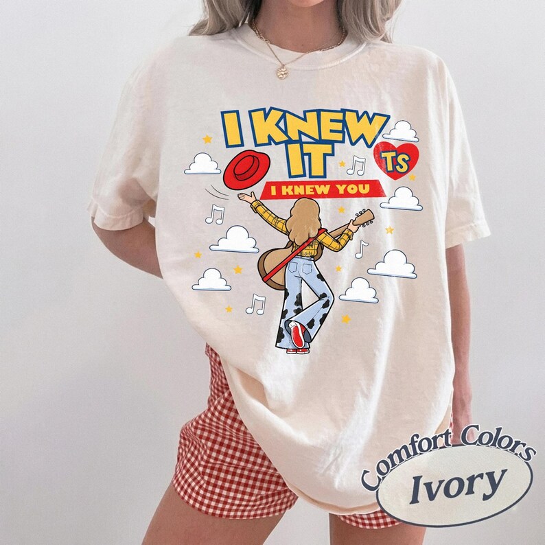 K&ouml;nnte beinhalten: Ein elfenbeinfarbenes T-Shirt mit einer Grafik einer Person mit langem blondem Haar, die Gitarre spielt und ein gelbes Karohemd sowie eine Hose mit Kuhfleckenmuster tr&auml;gt. Der Text lautet I KNEW IT und I KNEW YOU, erg&auml;nzt durch Wolken.