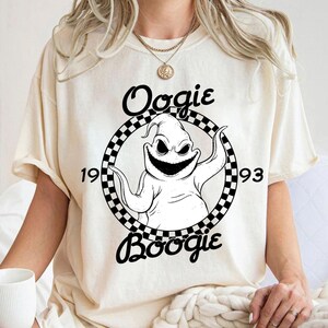 Pu&ograve; includere: Una t-shirt a maniche corte color crema con una grafica in bianco e nero di un personaggio all'interno di un cerchio a scacchi. Il testo "Oogie" appare sopra, "Boogie" sotto, e "19" e "93" ai lati del personaggio centrale.