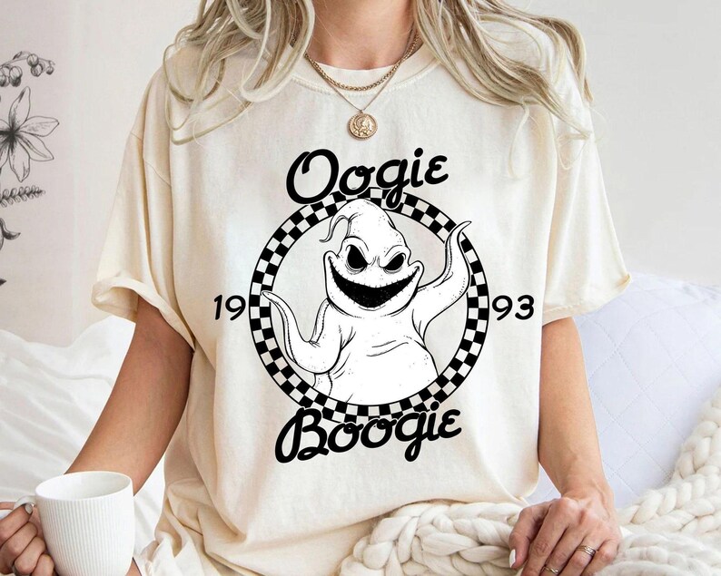Pu&ograve; includere: Una t-shirt a maniche corte color crema con una grafica in bianco e nero di un personaggio all'interno di un cerchio a scacchi. Il testo "Oogie" appare sopra, "Boogie" sotto, e "19" e "93" ai lati del personaggio centrale.