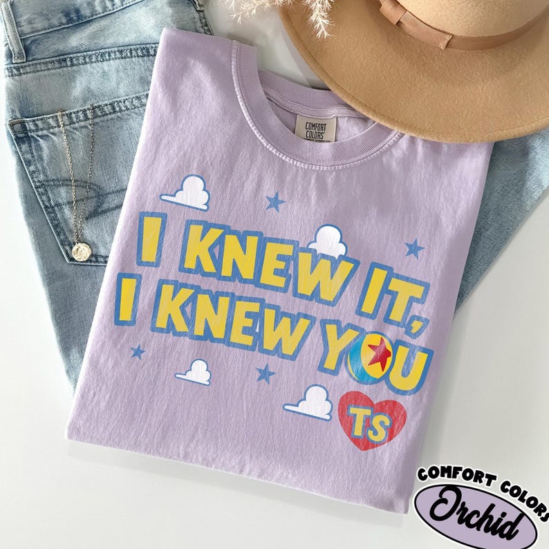 May include: A light purple t-shirt featuring the yellow text I KNEW IT, I KNEW YOU with blue outlines. The design includes white cloud shapes, blue stars, a colorful ball with a red star, and a red heart containing the yellow letters TS.