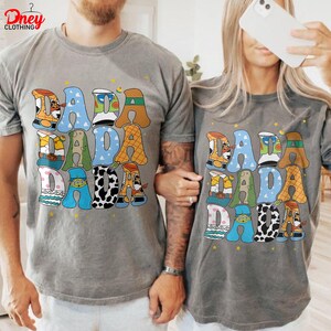 May include: Two gray t-shirts with the word "DADA" in large, colorful, cartoon-style letters. The letters are decorated with various patterns and characters. The shirts are worn by two people.