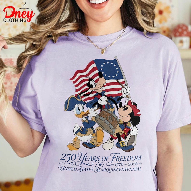 May include: A pale purple t-shirt with a graphic of Mickey Mouse, Donald Duck, and Goofy in patriotic outfits, holding a flag and drum. The shirt displays the text "250 Years of Freedom 1776-2026 United States Semiquincentennial."