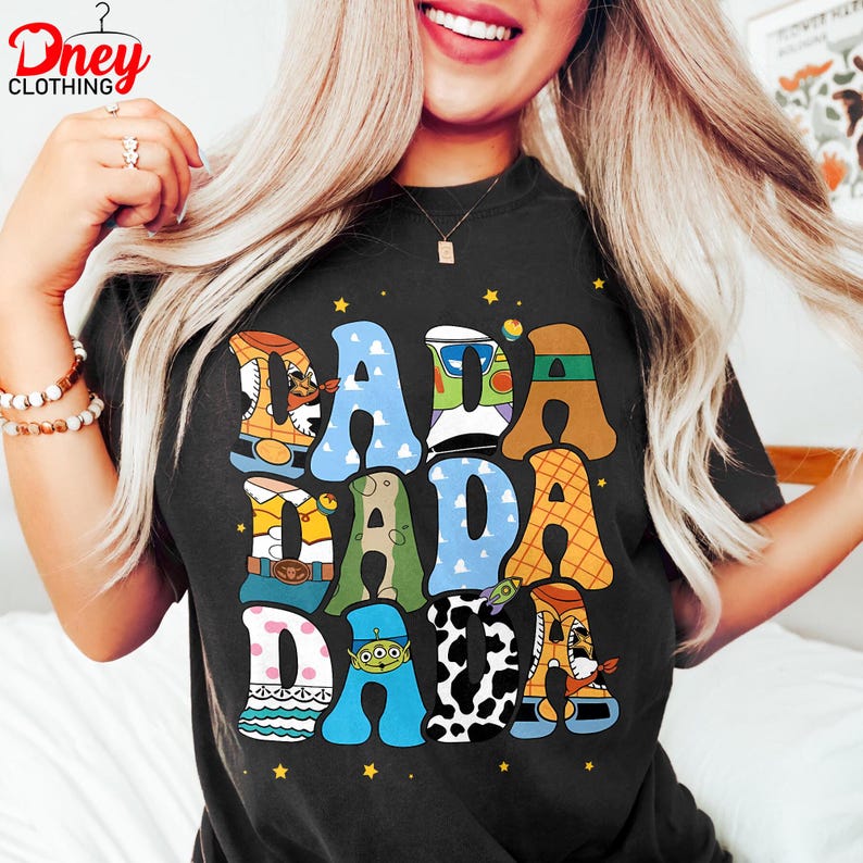 May include: Black t-shirt with the word "DADA" in colorful, cartoon-style letters. Each letter is designed with a different pattern and character theme, including a cowboy boot and a space alien. The shirt has a crew neck.