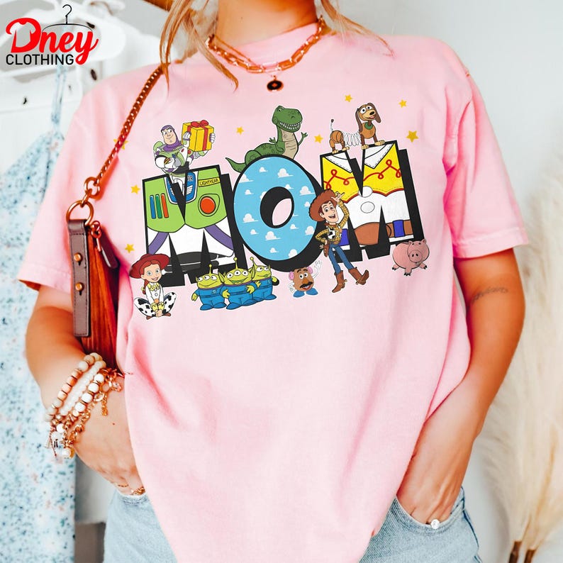 May include: Pale pink t-shirt featuring the word "MOM" in a colourful design with characters from the animated film Toy Story. The shirt includes images of Buzz Lightyear, Woody, and other characters.