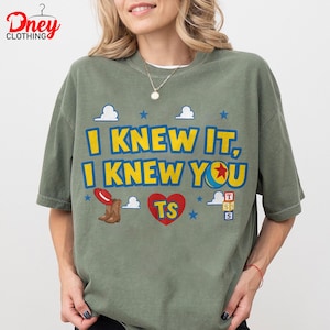Puede incluir: Una camiseta verde oliva con el texto amarillo I KNEW IT, I KNEW YOU, un coraz&oacute;n rojo con TS, un sombrero de vaquero rojo, botas marrones, una pelota colorida y estrellas azules. El dise&ntilde;o incluye nubes blancas y peque&ntilde;os cubos con letras.