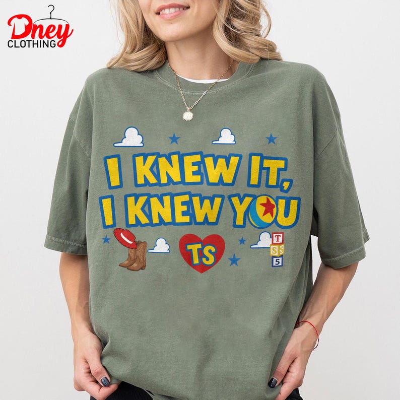 Puede incluir: Una camiseta verde oliva con el texto amarillo I KNEW IT, I KNEW YOU, un coraz&oacute;n rojo con TS, un sombrero de vaquero rojo, botas marrones, una pelota colorida y estrellas azules. El dise&ntilde;o incluye nubes blancas y peque&ntilde;os cubos con letras.