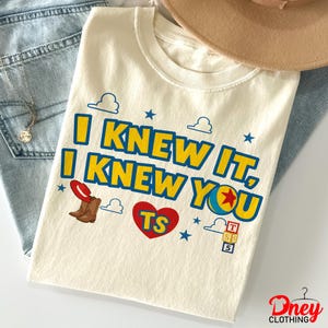 Puede incluir: Una camiseta de color crema con el texto en azul y amarillo I KNEW IT, I KNEW YOU. El dise&ntilde;o incluye un coraz&oacute;n rojo con las letras TS, un par de botas vaqueras marrones, una pelota colorida, bloques de letras apilados y nubes.