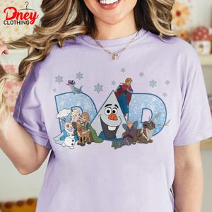 May include: A light purple t-shirt featuring the word "DAD" in a blue, snowflake-patterned design. The shirt showcases cartoon characters from a popular animated movie, including Olaf, Elsa, Anna, and Kristoff.