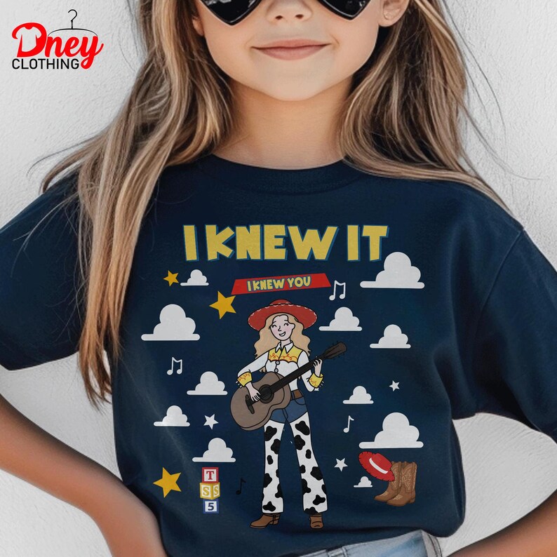 May include: A dark navy blue t-shirt featuring a graphic of a blonde cowgirl playing an acoustic guitar. The design includes the text I KNEW IT, white clouds, yellow stars, musical notes, a red hat, brown boots, and a block with the letters TSS 5.