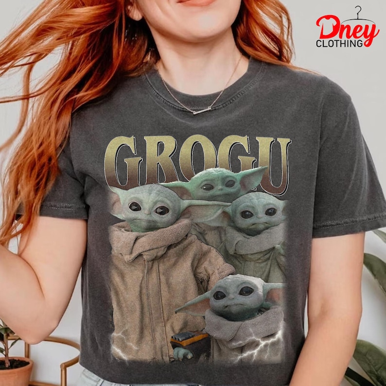 K&ouml;nnte beinhalten: Dunkelgraues T-Shirt mit dem goldenen Wort "GROGU" &uuml;ber einem Bild von Grogu, auch bekannt als Baby Yoda, aus der Star Wars-Serie. Das Bild zeigt Grogu in verschiedenen Posen.
