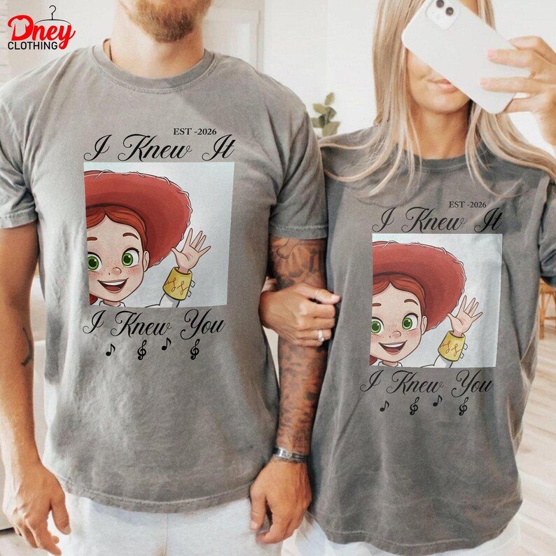 Op de afbeelding: Een grijs t-shirt met korte mouwen en een afbeelding van een stripfiguur met rood haar en een rode hoed, met gele manchetten. De tekst "I Knew It" staat boven de afbeelding, "EST-2026" bovenaan en "I Knew You" met muzieknoten eronder.
