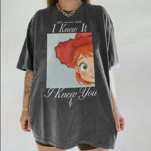 Puede incluir: Una camiseta gris carb&oacute;n de manga corta con el gr&aacute;fico de un personaje pelirrojo con sombrero de vaquero rojo. El texto dice EST 2026, I Knew It, e I Knew You, con un peque&ntilde;o s&iacute;mbolo de nota musical debajo del texto en la parte frontal.