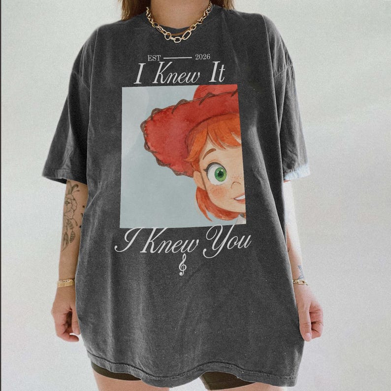 Puede incluir: Una camiseta gris carb&oacute;n de manga corta con el gr&aacute;fico de un personaje pelirrojo con sombrero de vaquero rojo. El texto dice EST 2026, I Knew It, e I Knew You, con un peque&ntilde;o s&iacute;mbolo de nota musical debajo del texto en la parte frontal.