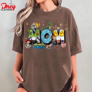 May include: Brown t-shirt with the word "MOM" in a colourful design featuring characters from the Toy Story film. The "O" in "MOM" is light blue with white polka dots. Yellow stars are scattered around the letters.