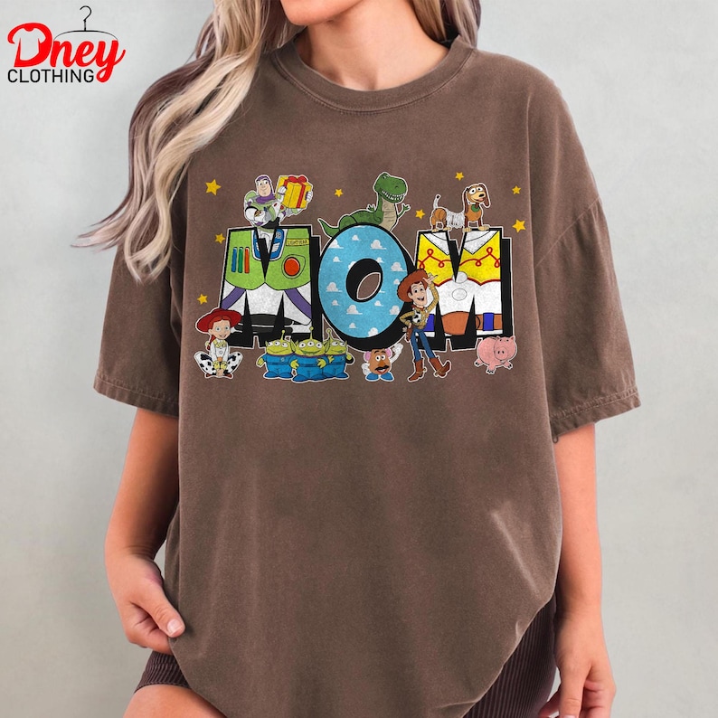 May include: Brown t-shirt with the word "MOM" in a colourful design featuring characters from the Toy Story film. The "O" in "MOM" is light blue with white polka dots. Yellow stars are scattered around the letters.
