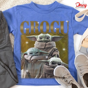 K&ouml;nnte beinhalten: Ein blaues T-Shirt mit einer Grafik von Grogu aus The Mandalorian. Das Wort "GROGU" ist goldfarben &uuml;ber dem Bild von Grogu aufgedruckt. Das Bild zeigt Grogu in einem braunen Umhang. Eine graue Hose und Schuhe liegen neben dem Shirt.