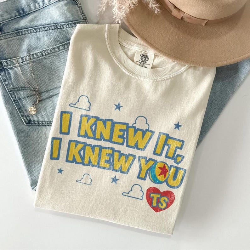 May include: A cream-colored short-sleeve t-shirt with the text I KNEW IT, I KNEW YOU printed in blue and yellow. The design includes blue stars, white cloud outlines, a red heart with the letters TS, and a colorful ball with a red star.
