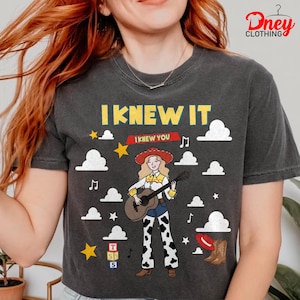 May include: A charcoal grey t-shirt featuring a graphic of a cartoon cowgirl with blonde hair playing an acoustic guitar. The text reads I KNEW IT and I KNEW YOU. The design includes white clouds, yellow stars, musical notes, alphabet blocks, and a brown boot.