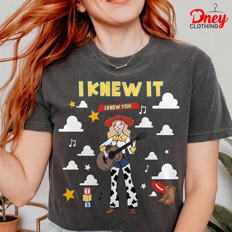 May include: A charcoal grey t-shirt featuring a graphic of a cartoon cowgirl with blonde hair playing an acoustic guitar. The text reads I KNEW IT and I KNEW YOU. The design includes white clouds, yellow stars, musical notes, alphabet blocks, and a brown boot.