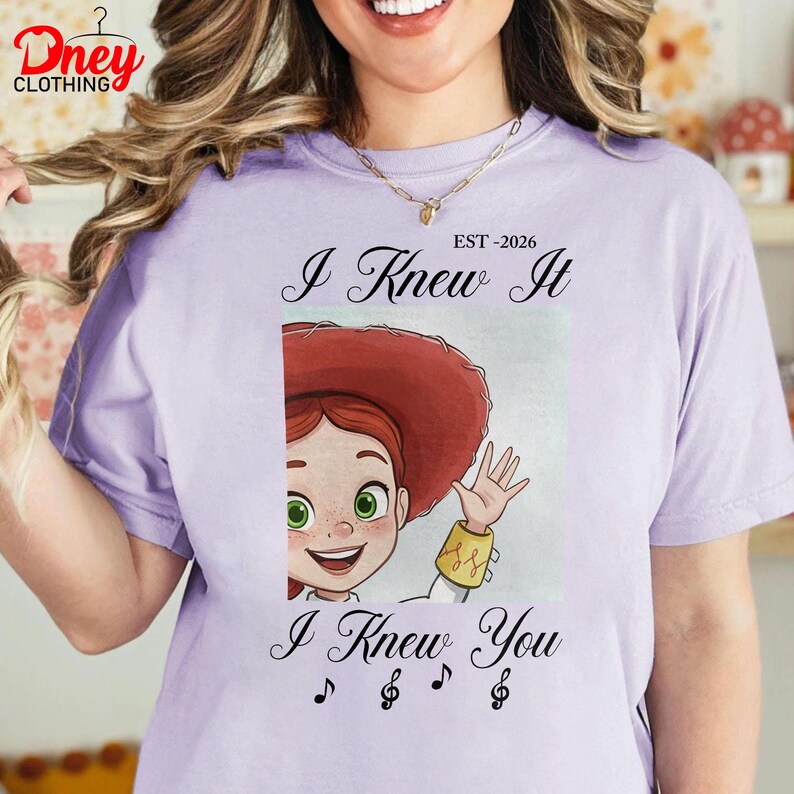 Op de afbeelding: Een lichtpaars t-shirt met een afbeelding van een stripfiguur met rood haar en een rode hoed die zwaait. De tekst luidt EST-2026, I Knew It en I Knew You, vergezeld door drie in zwarte inkt gedrukte muzieknootsymbolen.