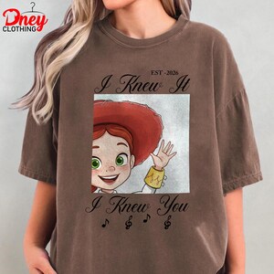Op de afbeelding: Een bruin t-shirt met korte mouwen en een afbeelding van een stripfiguur met rood haar en een rode hoed. De tekst luidt "I Knew It", "EST -2026" en "I Knew You", met muzieknootsymbolen onder de illustratie van het personage.