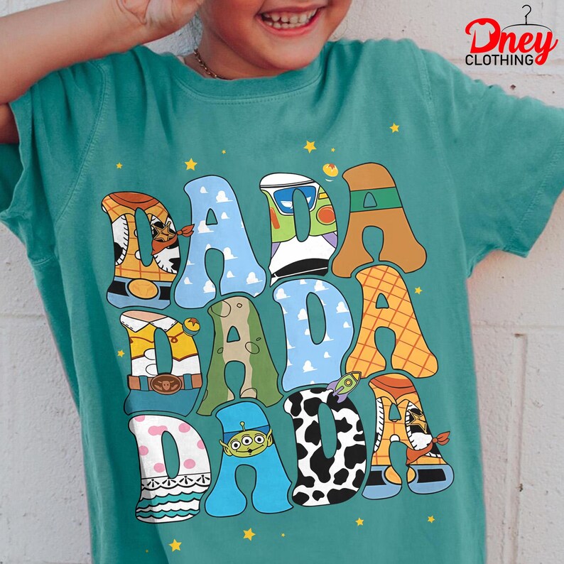 May include: Teal t-shirt with the word "DADA" in colorful, cartoon-style letters. Each letter is designed with different patterns and character themes, including a cowboy hat, clouds, and a cow print. The shirt has a crew neck and short sleeves.