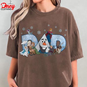May include: Brown t-shirt featuring the word "DAD" in blue, with snowflakes and characters from the movie Frozen. The shirt has a crew neck and short sleeves. The brand "Dney Clothing" is printed on the top left.