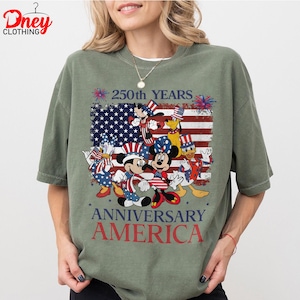Puede incluir: Camiseta verde oliva con un dise&ntilde;o de Mickey y Minnie Mouse, el Pato Donald, Goofy y Pluto con atuendos patri&oacute;ticos. El dise&ntilde;o incluye una bandera estadounidense y el texto "250th YEARS ANNIVERSARY AMERICA."