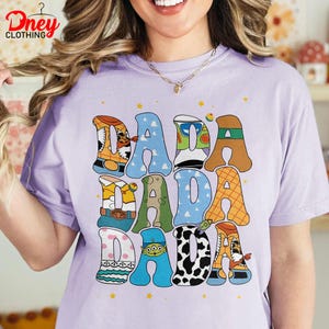 May include: A light purple t-shirt with the word "DADA" repeated three times in a playful, colorful design. Each letter is decorated with a different pattern, including a cow print, a toy character, and a blue sky.