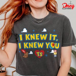 Puede incluir: Una camiseta gris carb&oacute;n de manga corta con el texto en amarillo y azul I KNEW IT, I KNEW YOU. El dise&ntilde;o incluye un sombrero de vaquero rojo, una bota marr&oacute;n, un coraz&oacute;n rojo con las letras TS, una pelota azul y amarilla y nubes blancas.