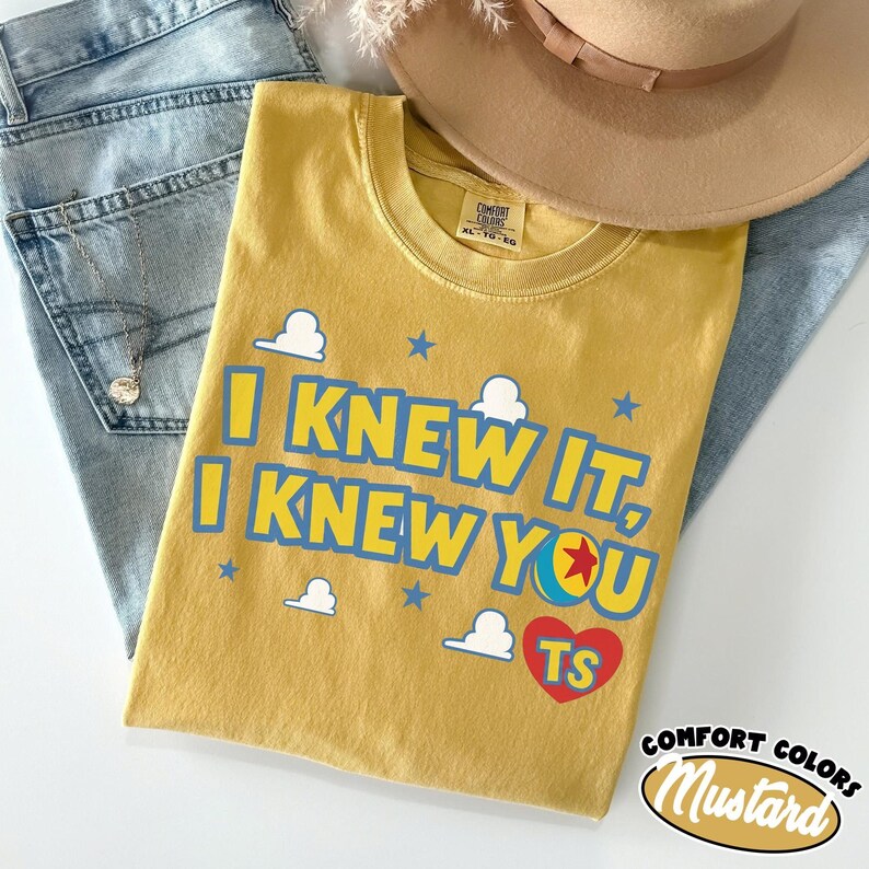 May include: A mustard yellow t-shirt featuring the blue text I KNEW IT, I KNEW YOU with a red heart containing the letters TS and a small ball graphic. The shirt has a crew neckline and a Comfort Colors label. The design includes white clouds and blue stars.