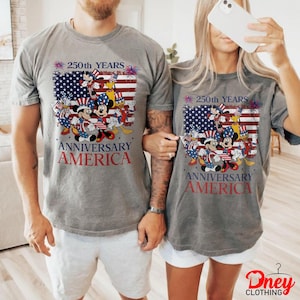 Puede incluir: Dos camisetas grises con un gr&aacute;fico que celebra el 250 aniversario de Am&eacute;rica. El dise&ntilde;o presenta personajes de Disney con atuendo patri&oacute;tico, sobre un fondo de la bandera estadounidense. El texto dice '250th YEARS ANNIVERSARY AMERICA'.