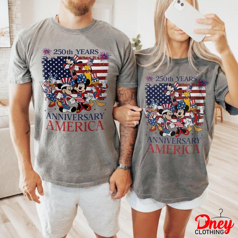 Puede incluir: Dos camisetas grises con un gr&aacute;fico que celebra el 250 aniversario de Am&eacute;rica. El dise&ntilde;o presenta personajes de Disney con atuendo patri&oacute;tico, sobre un fondo de la bandera estadounidense. El texto dice '250th YEARS ANNIVERSARY AMERICA'.