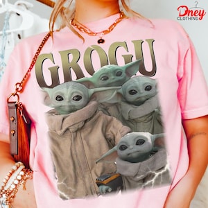 K&ouml;nnte beinhalten: Hellrosa T-Shirt mit einem Grogu-Motiv aus The Mandalorian. Das Design zeigt mehrere Bilder von Grogu mit dem Wort "GROGU" in gro&szlig;en, goldenen Buchstaben. Das T-Shirt hat eine hellrosa Farbe.