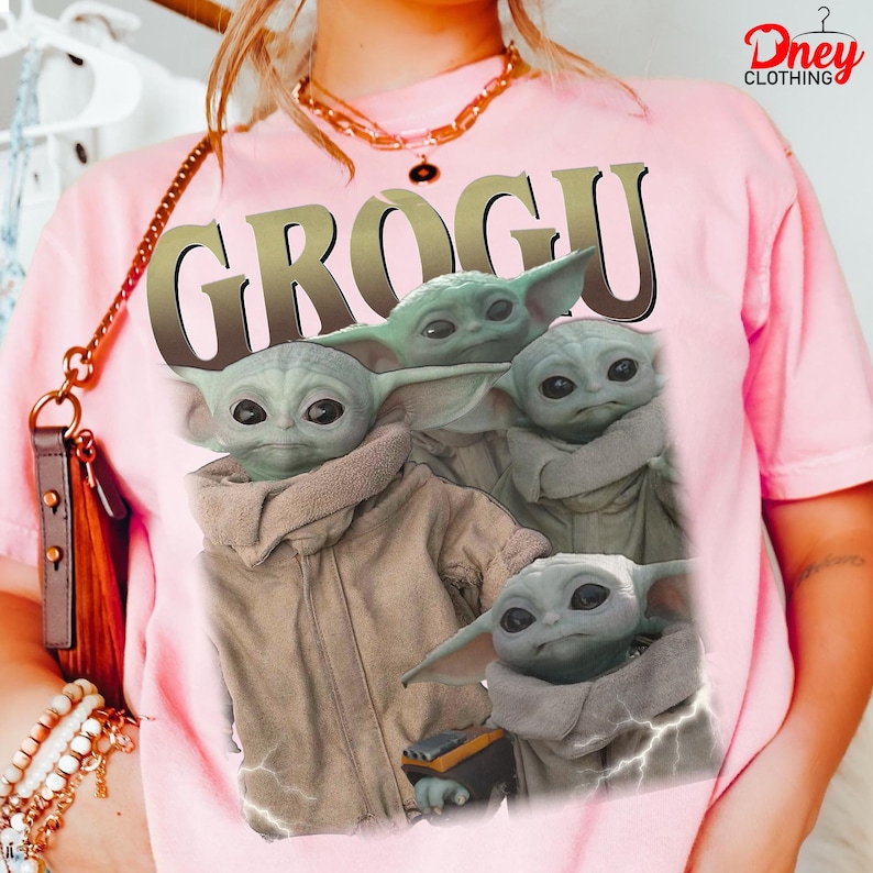 K&ouml;nnte beinhalten: Hellrosa T-Shirt mit einem Grogu-Motiv aus The Mandalorian. Das Design zeigt mehrere Bilder von Grogu mit dem Wort "GROGU" in gro&szlig;en, goldenen Buchstaben. Das T-Shirt hat eine hellrosa Farbe.